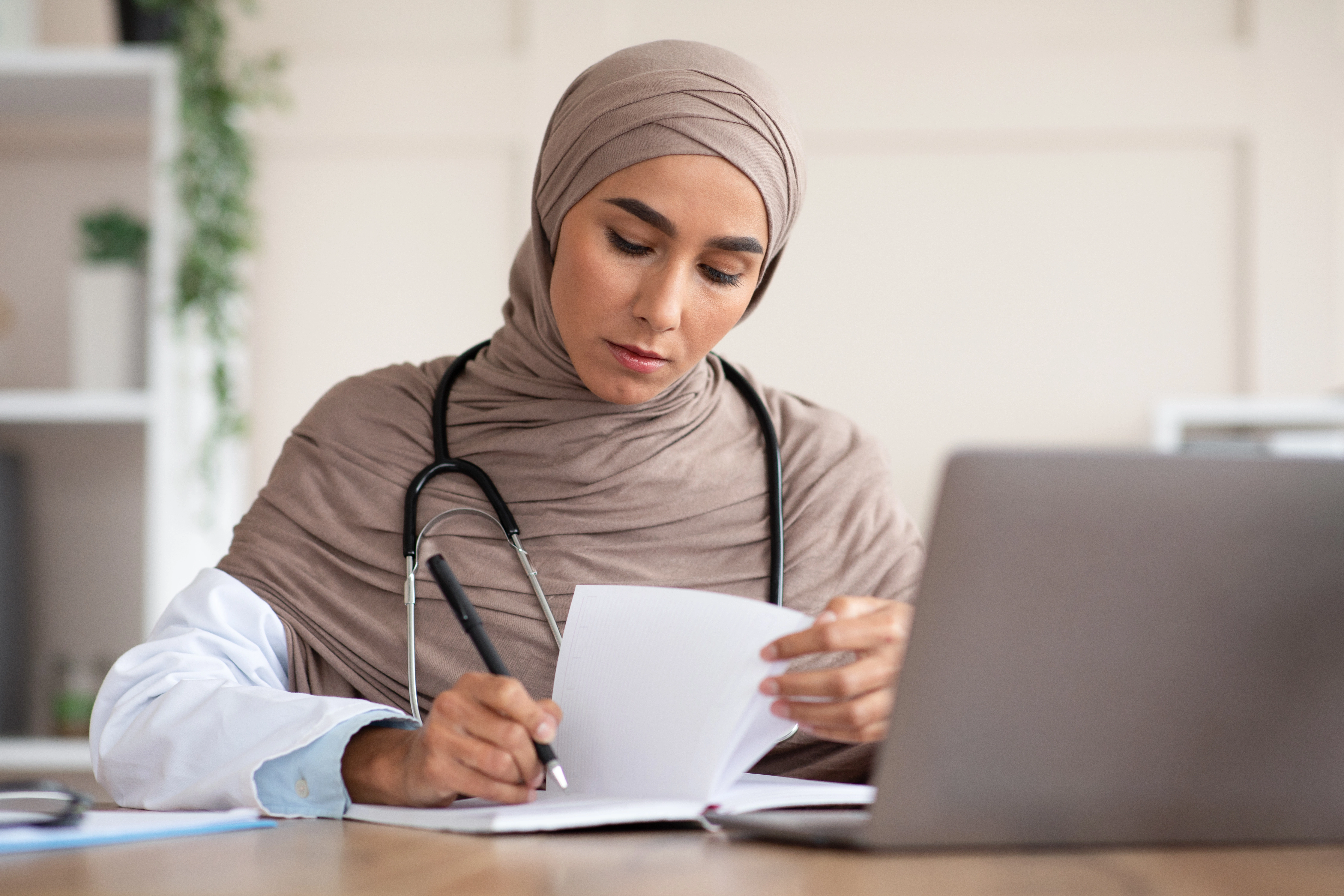 Concentrated young woman medical professional taking notes with laptop